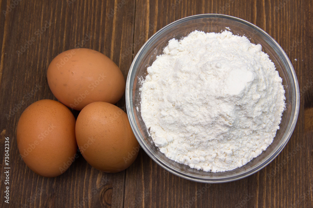 Eggs and flour in bowl on wooden table seen from above