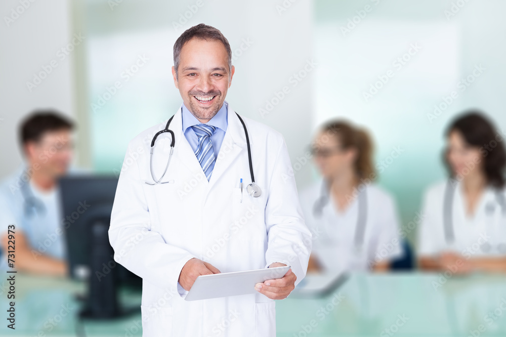 Doctor Holding Digital Tablet Against Team In Meeting Room Doctor Holding Digital Tablet Against Team In Meeting Room