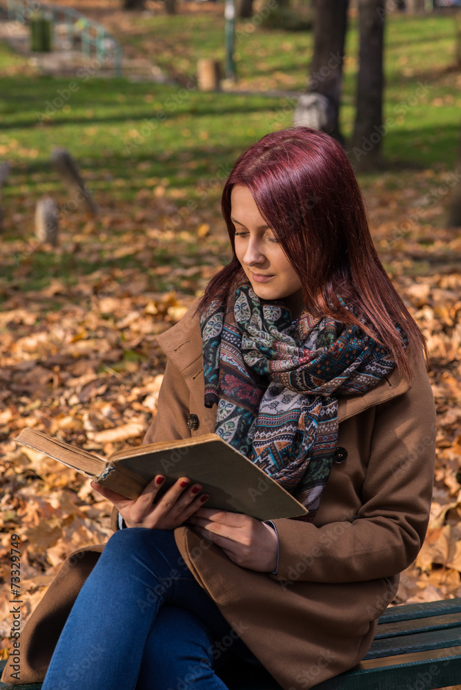 Obraz premium redhead girl sitting on bench in park and reading book