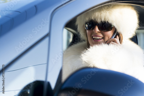 Woman in car wearing white fur coat on the phone smiling