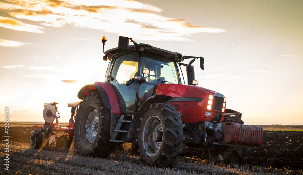 Obraz premium Farmer plowing stubble field with red tractor at sunset