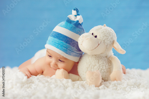 Cmile baby in hat, hugging toy on a white bedspread, on a blue b