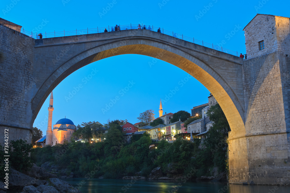 Illuminated old Ottoman bridge in Mostar at dusk with buildings of Čaršija and the mosque in the background, Bosnia and Herzegovina