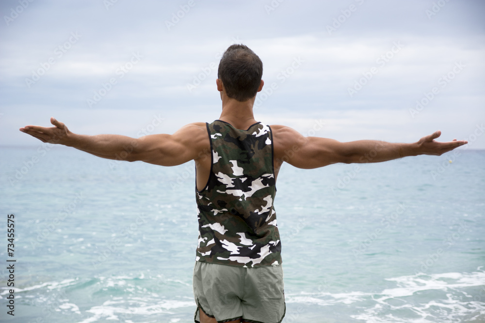 Muscular young man on the beach seen from the back, with arms op Muscular young man on the beach seen from the back, with arms op