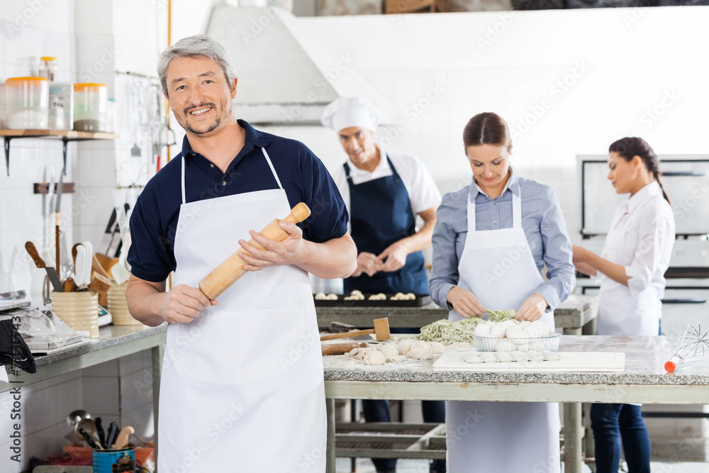 Confident Male Chef Holding Rolling Pin While Colleagues Prepari Confident Male Chef Holding Rolling Pin While Colleagues Prepari