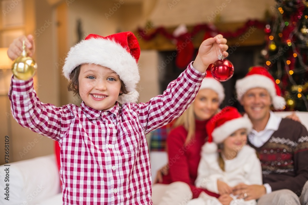 Son wearing santa hat holding baubles in front of his family Son wearing santa hat holding baubles in front of his family