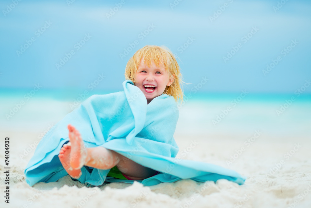 Portrait of laughing baby girl wrapped in towel sitting on beach