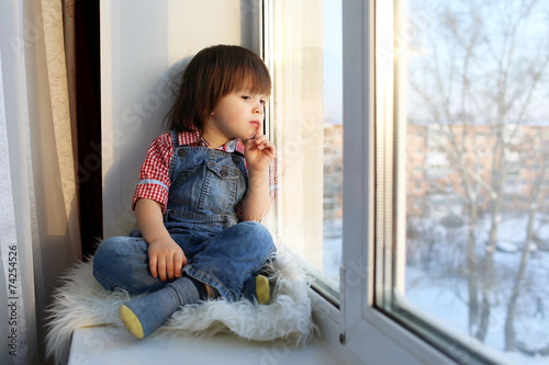 Lovely little boy sits on sill and looks out of window in winter