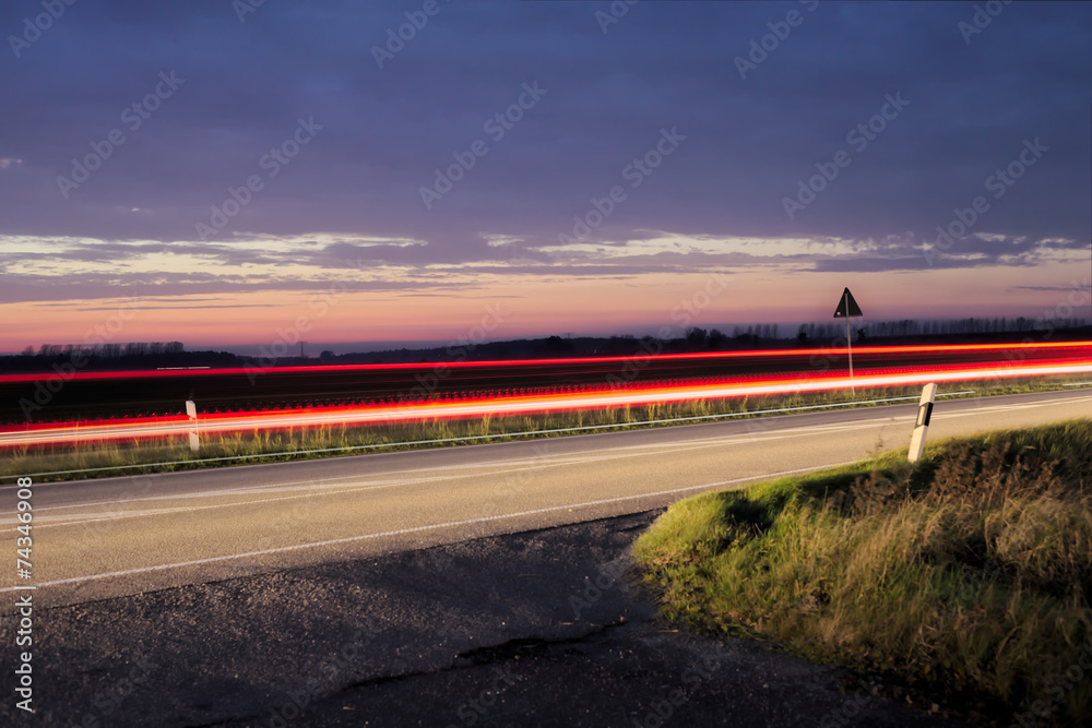 Fototapeta premium Sunset over a federal road (Bundesstrasse) near Greifswald, Mecklenburg-Vorpommern, Germany. Long exposure makes the movement of car lights visible