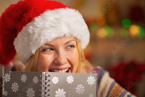 Portrait of smiling girl in santa hat hiding behind notpad photo