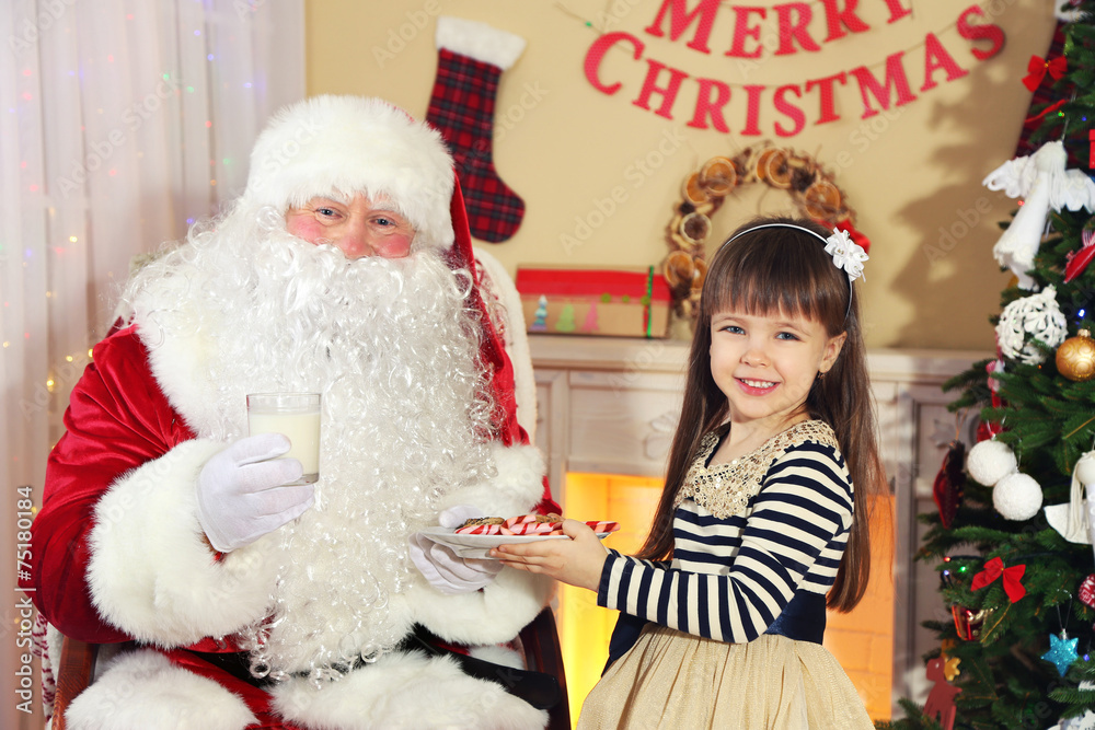 Obraz premium Little cute girl giving cookies and milk to Santa Claus near