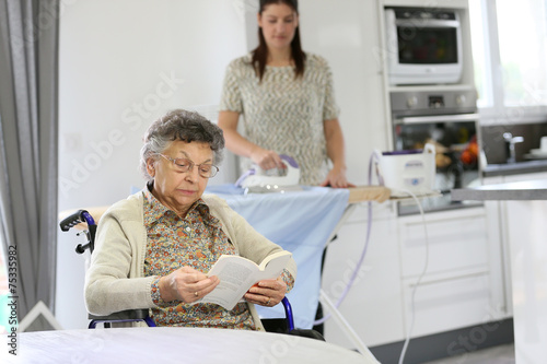 Quadro su tela Elderly woman reading book while home helper irons laundry