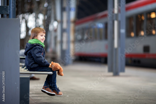 Cute boy, sitting on a bench with teddy bear, looking at a train