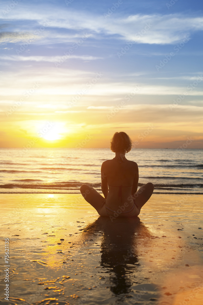 Young girl sitting meditation on the ocean during an sunset.