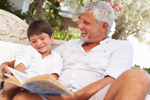 Grandfather And Grandson Sitting In Garden Reading Book