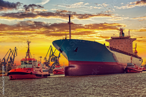 Big ship with escorting tugs leaving port at sunset.