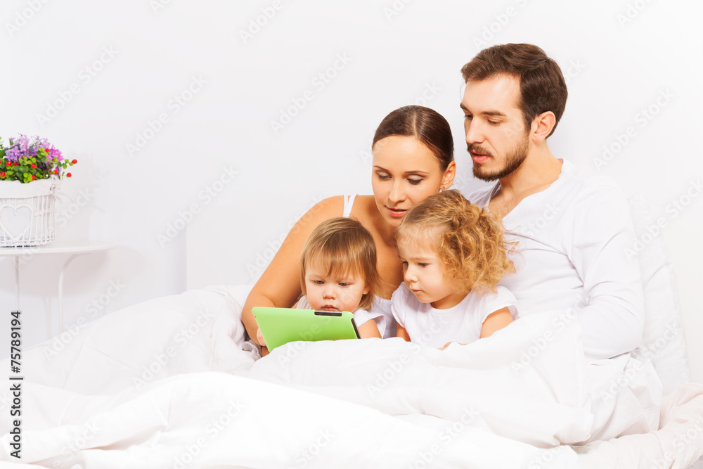 Parents and two cute kids play with tablet on bed