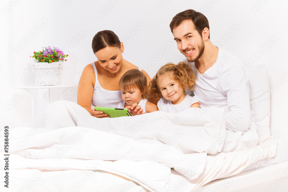 Parents and two kids play with tablet on white bed