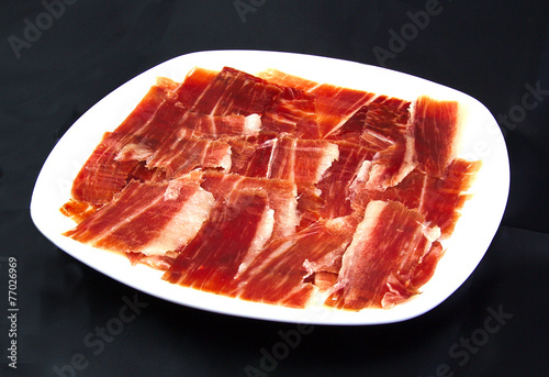 Serrano ham slices on a white dish over black wackground. Jabugo