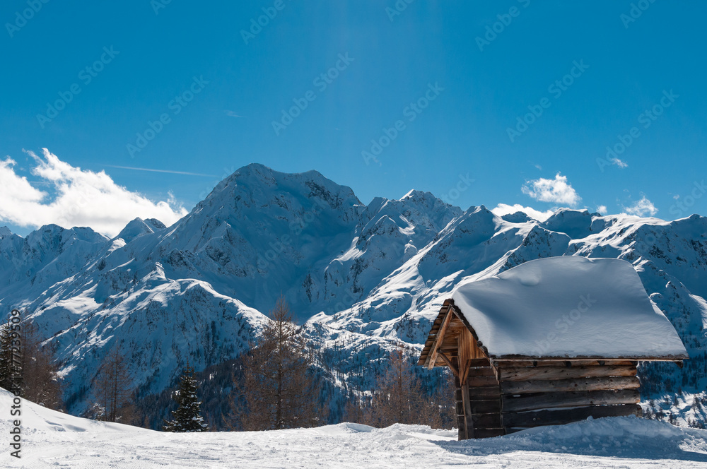 Wooden hut covered with snow in front of high mountains.