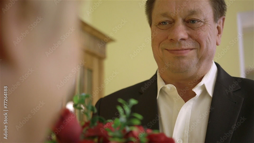 Middle-aged man giving woman bouquet of red roses, indoors.