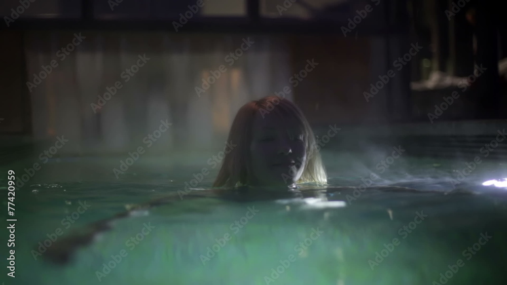 Shot of a pretty young woman in blue bikini that swimms towards camera in the outdoor pool in the night Shot of a pretty young woman in blue bikini that swimms towards camera in the outdoor pool in the night