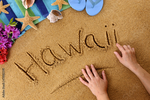 Child hands drawing the word Hawaii written in sand on a beach with towel flip flops seashells Hawaiian summer vacation holiday photo