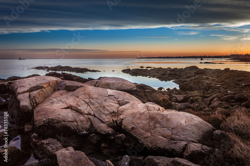 Boulders On The Shore At Sunset With Lighthouse In The Distance