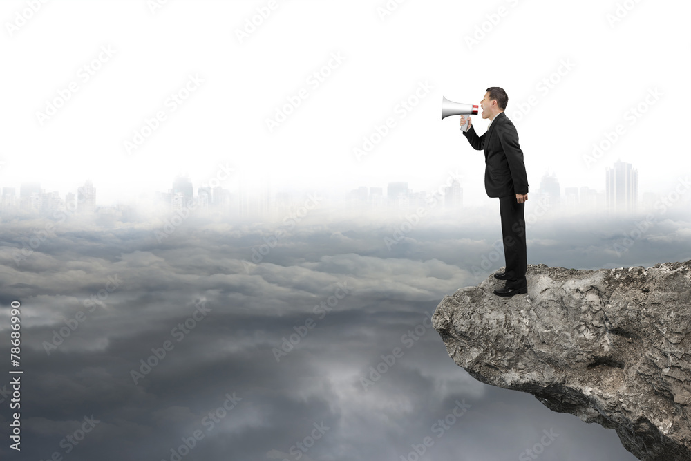 Businessman using megaphone yelling on cliff with gray cloudy ci