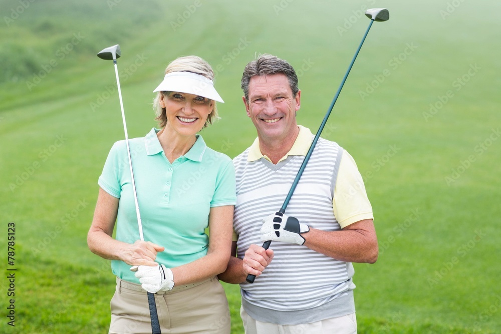 Golfing couple smiling at camera holding clubs - obrazy, fototapety, plakaty 