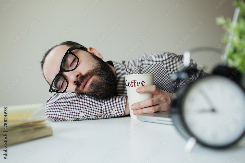 Man sleeps in office on table over laptop with coffee in hand - obrazy, fototapety, plakaty 