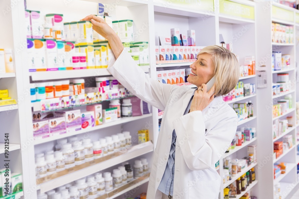 Smiling pharmacist phoning and taking medicine from shelf