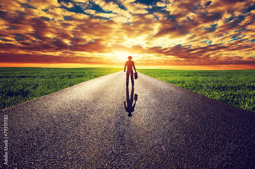 Man with suitcase and hat on long straight road to sunset sun