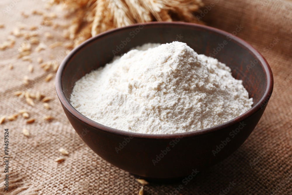 Flour in bowl with ears and grains on sackcloth background