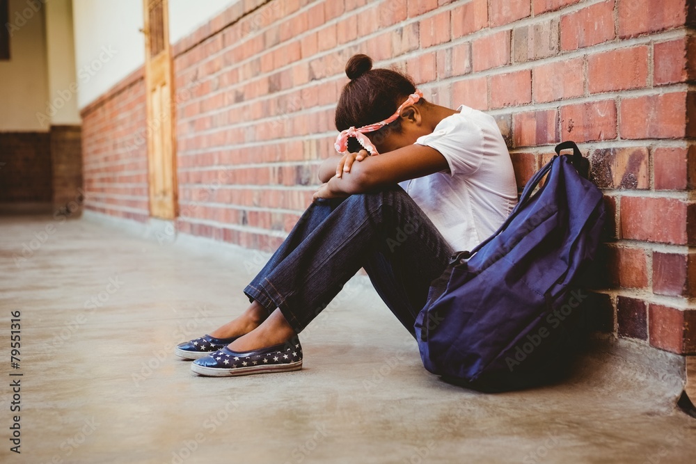 Tensed mixed race girl sitting against brick wall in school corridor - obrazy, fototapety, plakaty 