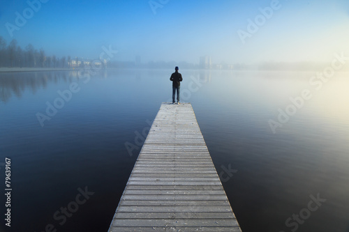 Man standing alone on a jetty, looking over a foggy lake.