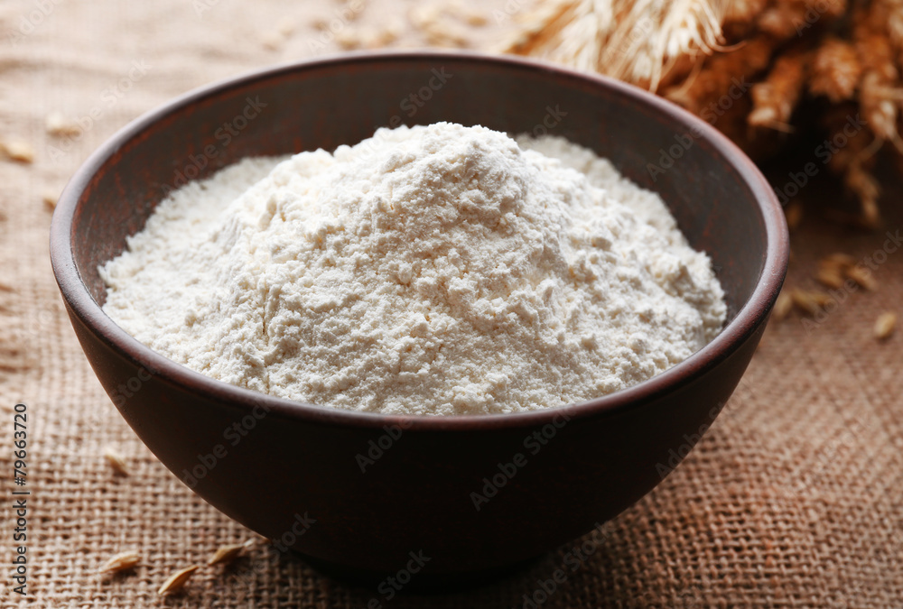 Flour in bowl with ears and grains on sackcloth background