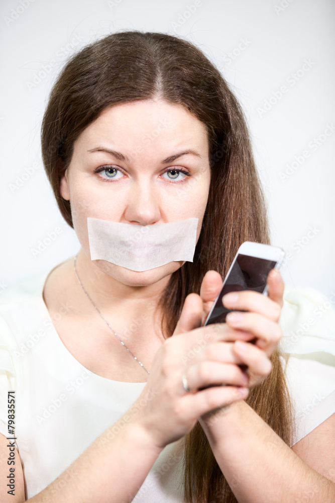 Caucasian woman with mouth sealed tape holds cell phone in hands Caucasian woman with mouth sealed tape holds cell phone in hands
