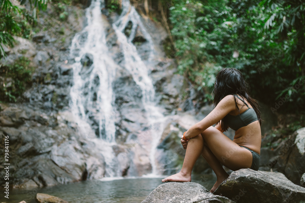 Asian tourist wearing a bikini sitting on a rock by idyllic natural waterfall in Khao Lak, Phang Nga province, Southern Thailand