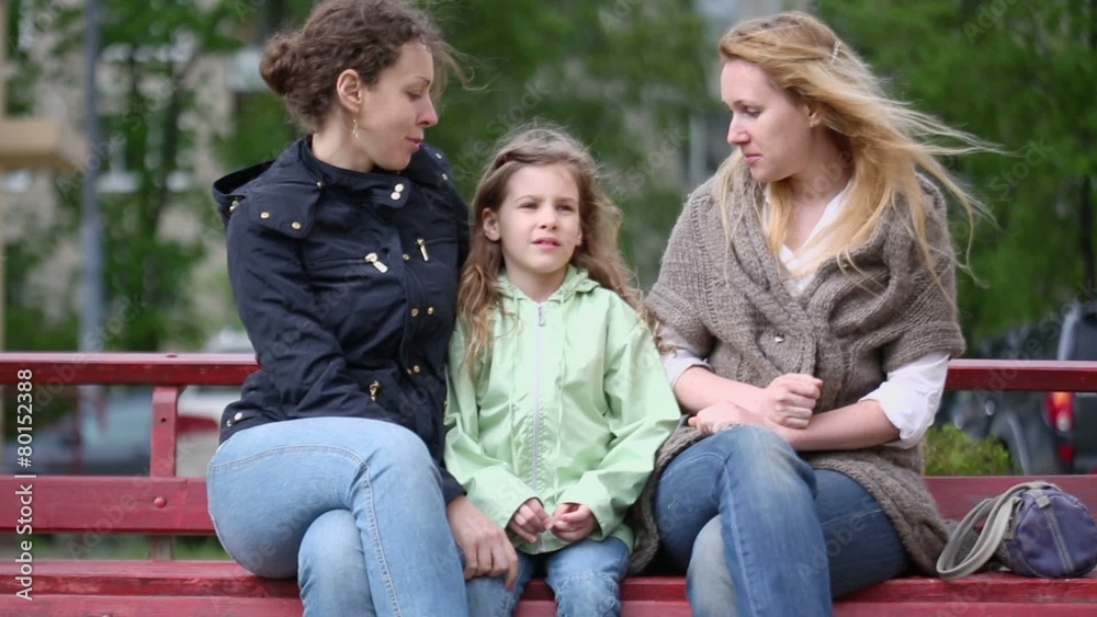 Two young women and little girl talk during sit on bench at yard Two young women and little girl talk during sit on bench at yard