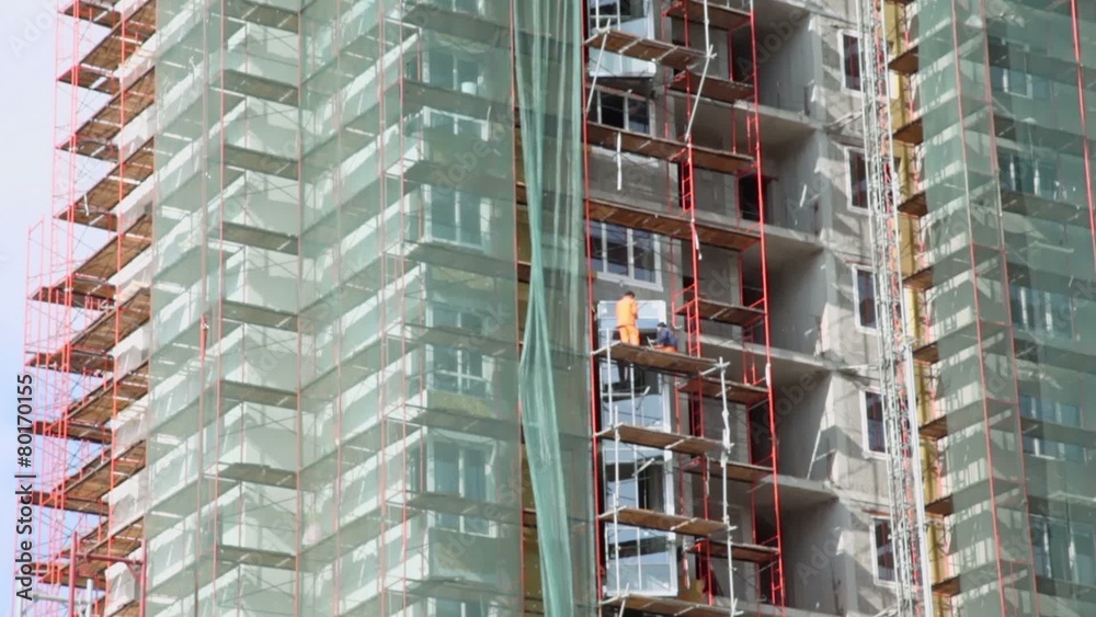 Workers stand on scaffold and set windows at construction site