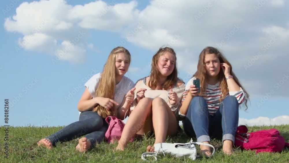 Three girls with bags sways and sing when listen music from cell phones on grass hill at sunny summer day Three girls with bags sways and sing when listen music from cell phones on grass hill at sunny summer day