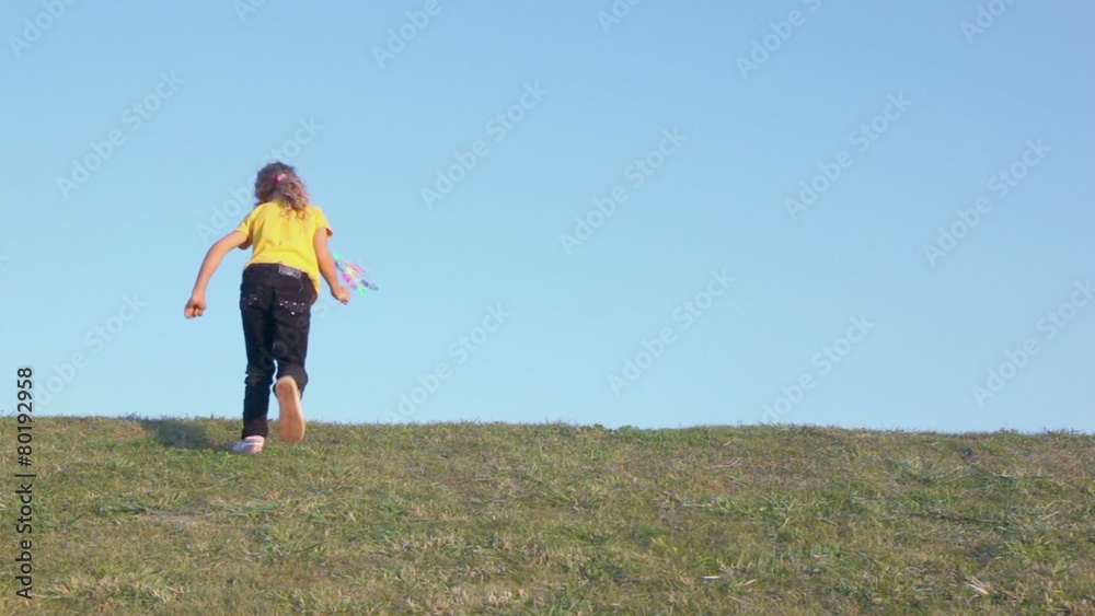 Little girl holds windmill toy and runs away on grass hill Little girl holds windmill toy and runs away on grass hill