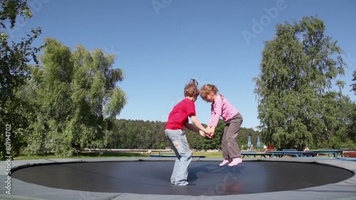 Two kids boy with little girl hold hands and try to jump on trampoline in park at summer day photo