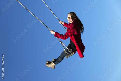 Happy young lady with a red coat swinging in blue sky