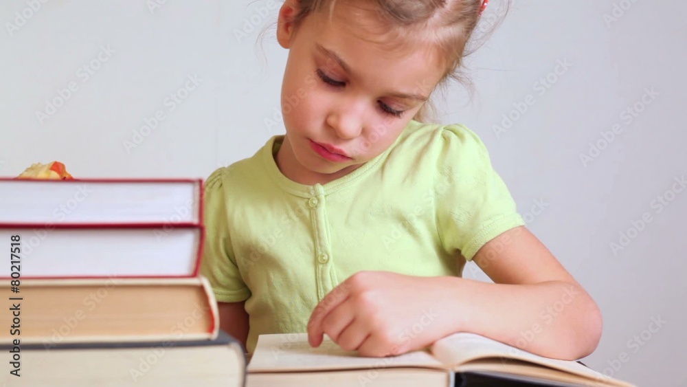 little girl near the pile of books reads book and eats apple little girl near the pile of books reads book and eats apple