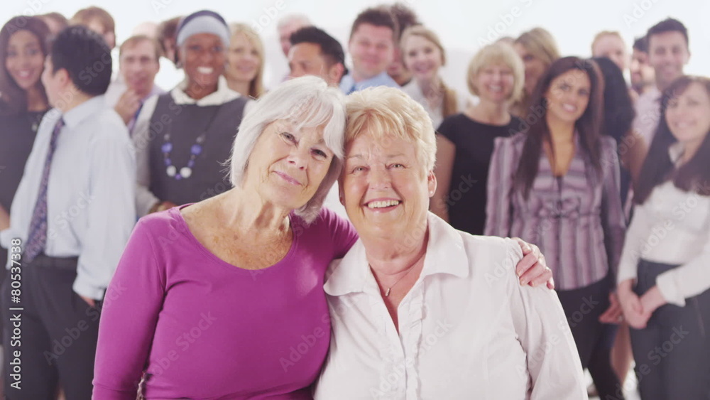 2 cheerful mature ladies standing in front of large diverse group of people 2 cheerful mature ladies standing in front of large diverse group of people