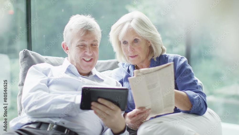 Attractive mature couple relaxing at home with a newspaper and computer tablet Attractive mature couple relaxing at home with a newspaper and computer tablet