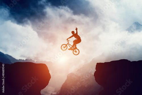 Man jumping on bmx bike over precipice in mountains at sunset.