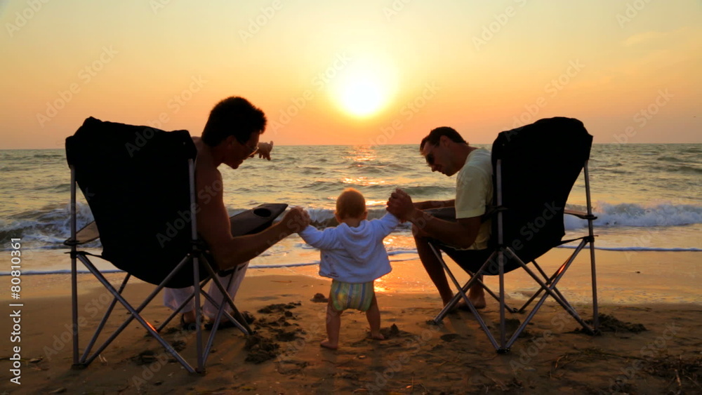 Two Men In Outdoor Chairs And Baby On The Beach At Sunset Two Men In Outdoor Chairs And Baby On The Beach At Sunset